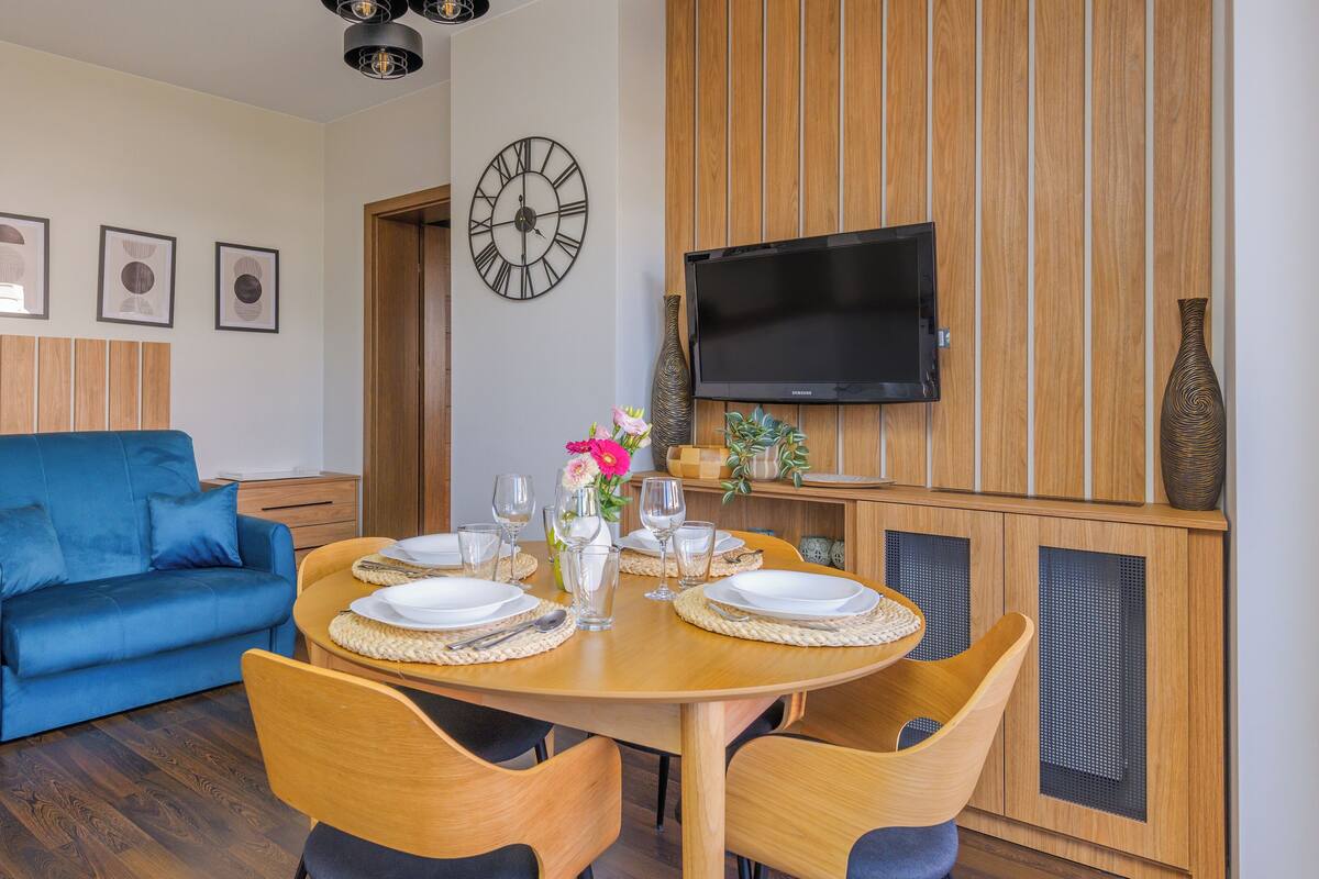 A modern living room with a blue sofa, a wooden dining table set for four, and a large wall clock.
