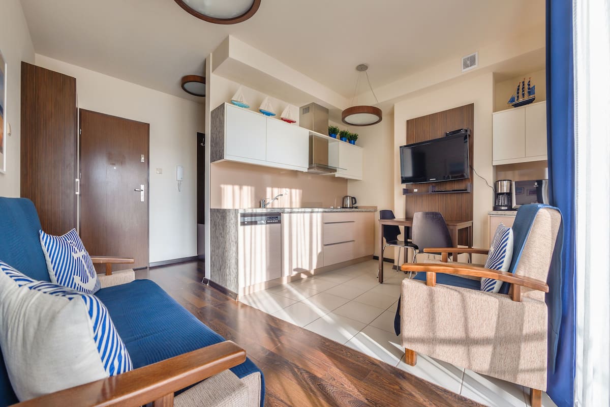 A modern living room with a sofa and armchairs, a kitchen with a dishwasher and microwave, and a television.