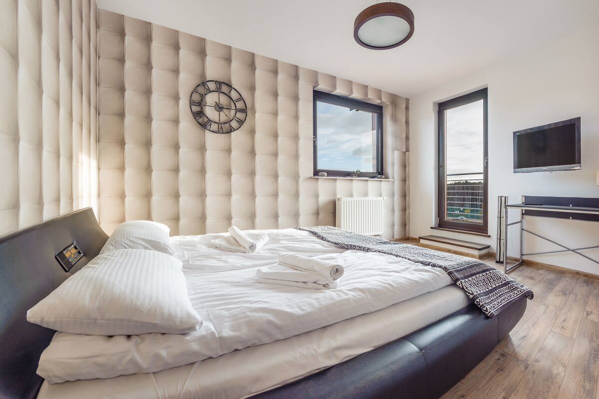 A modern bedroom with a large bed, a wall clock, a window, and a TV.