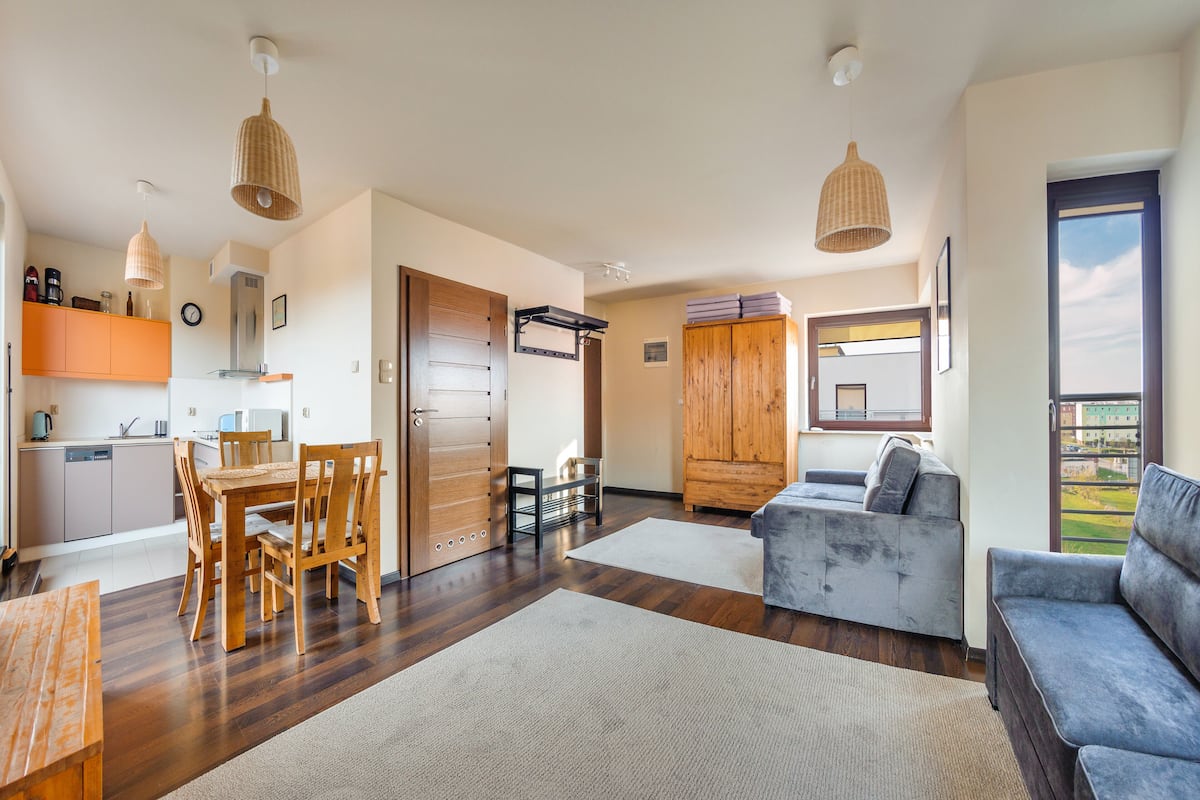 A modern living room with a grey sofa, wooden dining table, and chairs. There is a kitchen area with cabinets and a microwave. The room has wooden flooring and large windows.