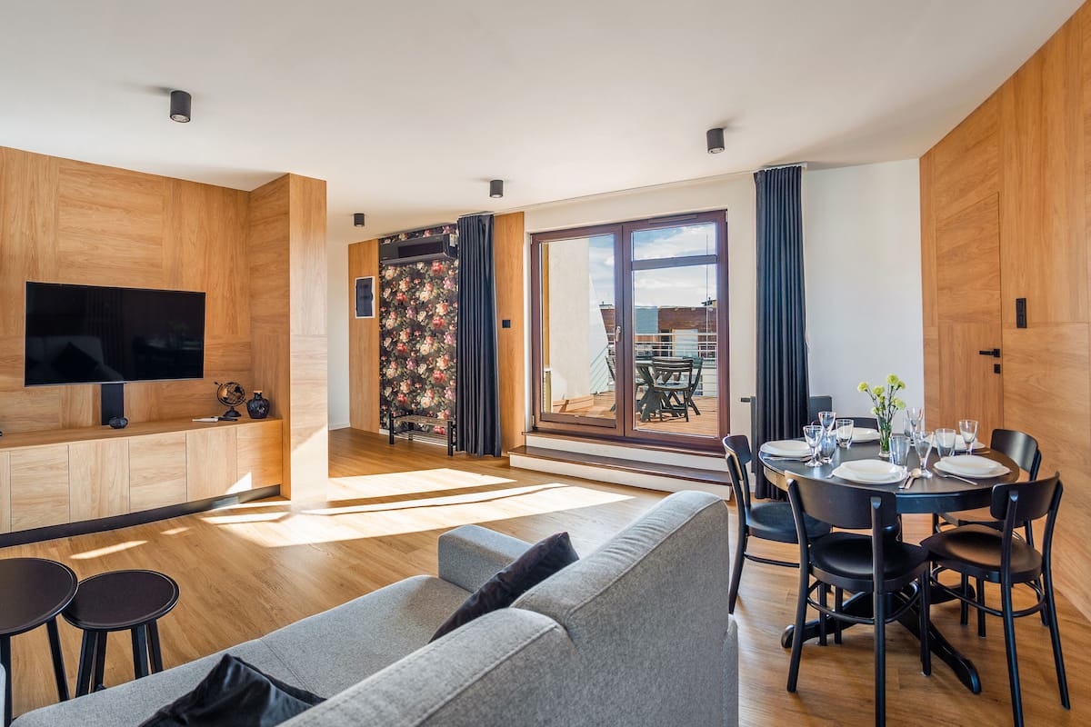 A modern living room with a grey sofa, a dining table set for four, and a TV mounted on a wooden wall.