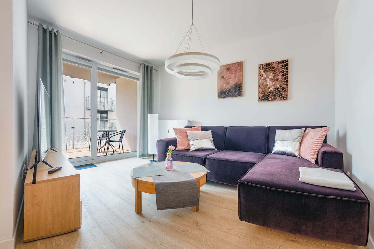 Uma sala de estar moderna com um sofá roxo, uma mesa de centro de madeira e uma porta de vidro que dá para uma varanda externa.