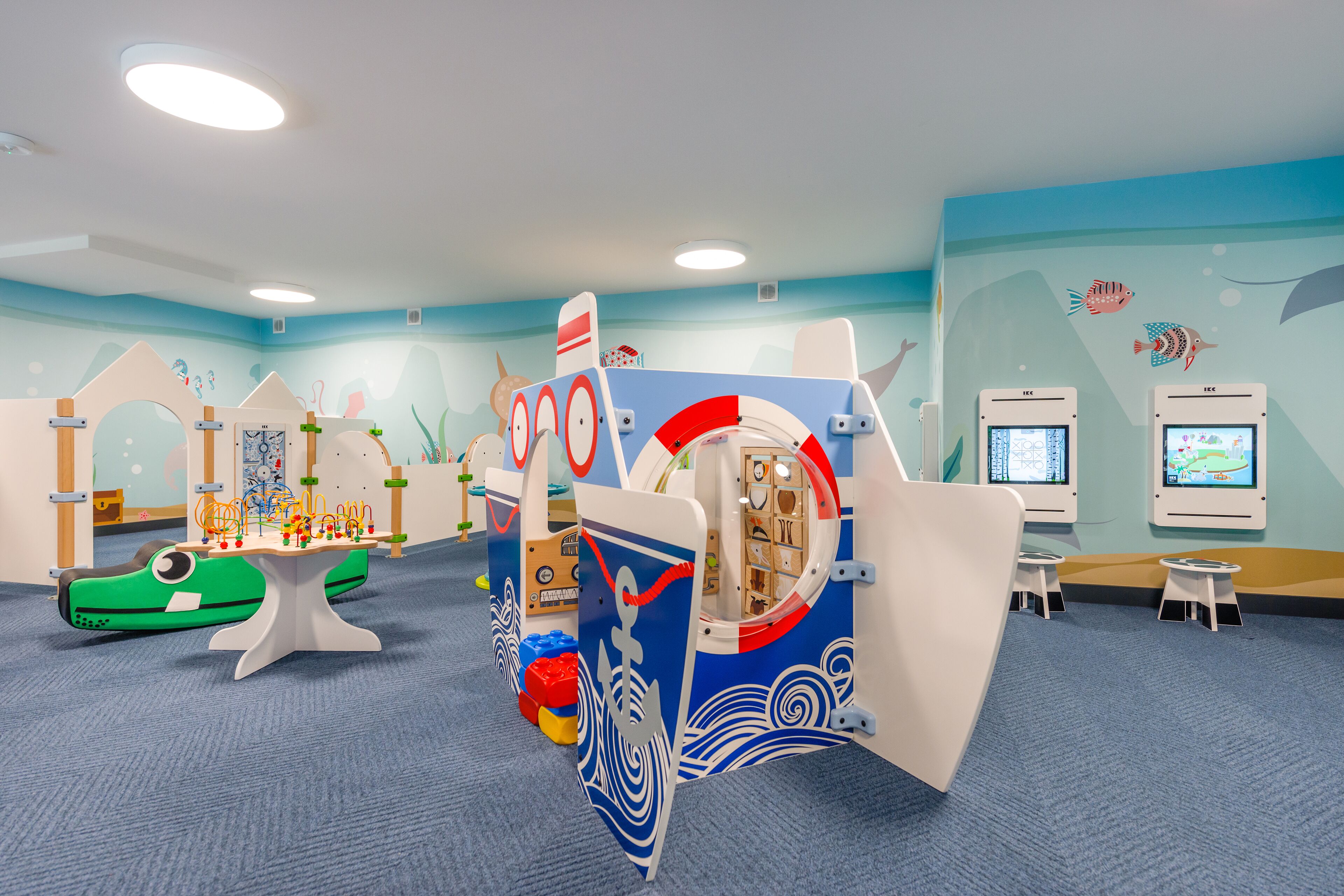 Ein Kinderspielbereich mit maritimen Elementen, darunter eine schiffsförmige Spielkonstruktion und Lernbildschirme.