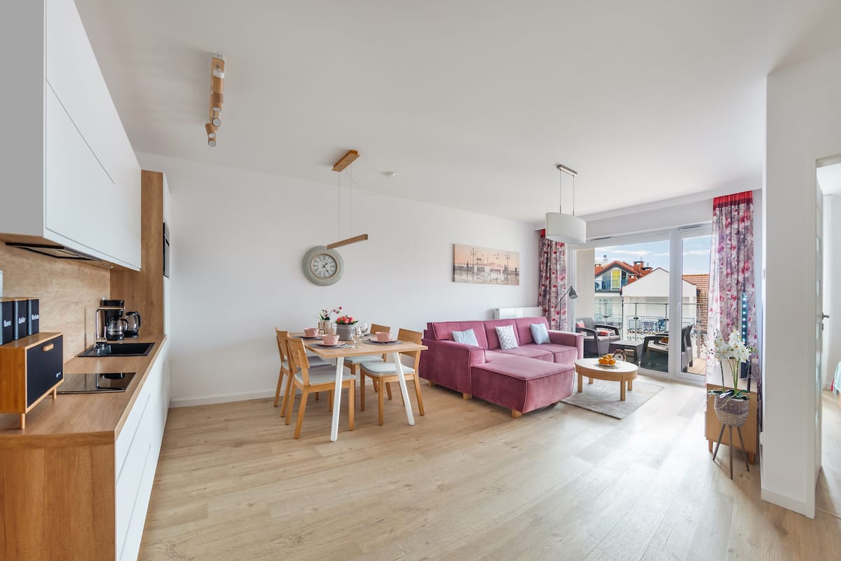 Uma sala de estar moderna com uma mesa de jantar de madeira, um sofá rosa e vista para a paisagem urbana através de grandes janelas.