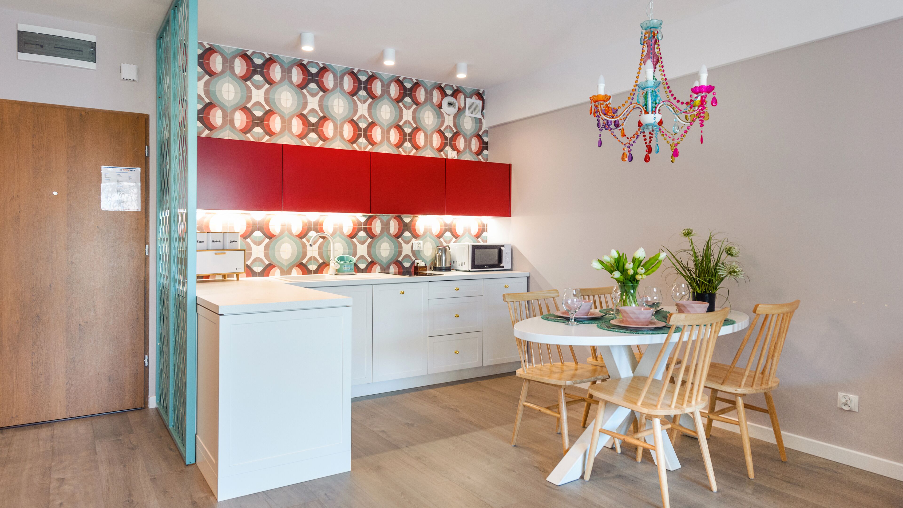 Una cocina moderna con una isla central, armarios blancos y salpicadero rojo. Se observa una mesa de comedor con sillas y una lámpara de araña colgante.