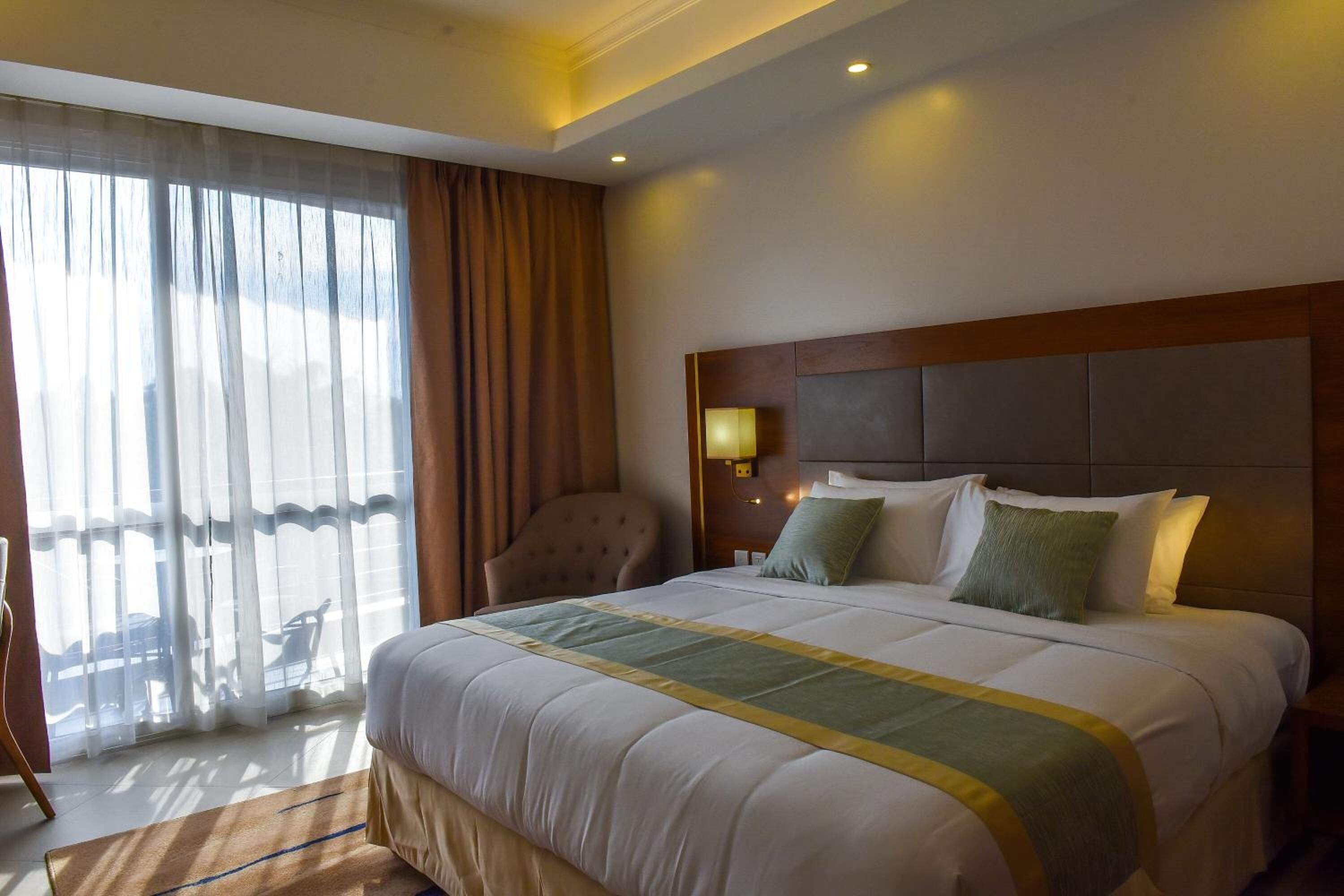 A hotel room with a large bed, a headboard, a nightstand with a lamp, a chair, and a view of the outdoors through sheer curtains.