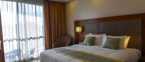 A hotel room with a large bed, a headboard, a nightstand with a lamp, a chair, and a view of the outdoors through sheer curtains.