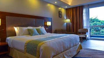 Een hotelkamer met een groot bed, een houten hoofdbord, twee bedlampen, een stoel, een bijzettafeltje en uitzicht op groen door een raam.