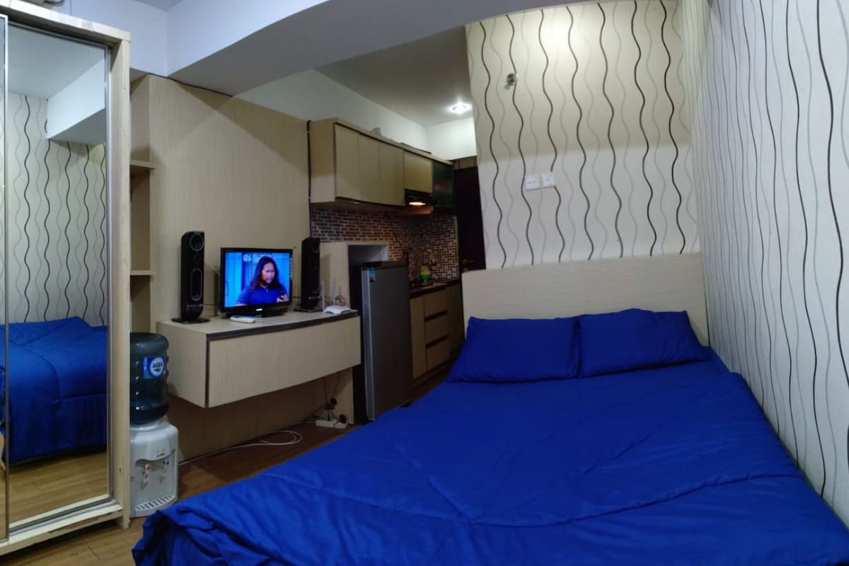 A bedroom with a bed, a TV, a water dispenser, and a kitchenette.