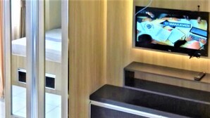 A hotel room with a large flat-screen TV mounted on the wall, a dark wooden cabinet below, and a sliding glass door leading to a balcony. - The Jarrdin Apartment by Berdikari Group (Bandung)