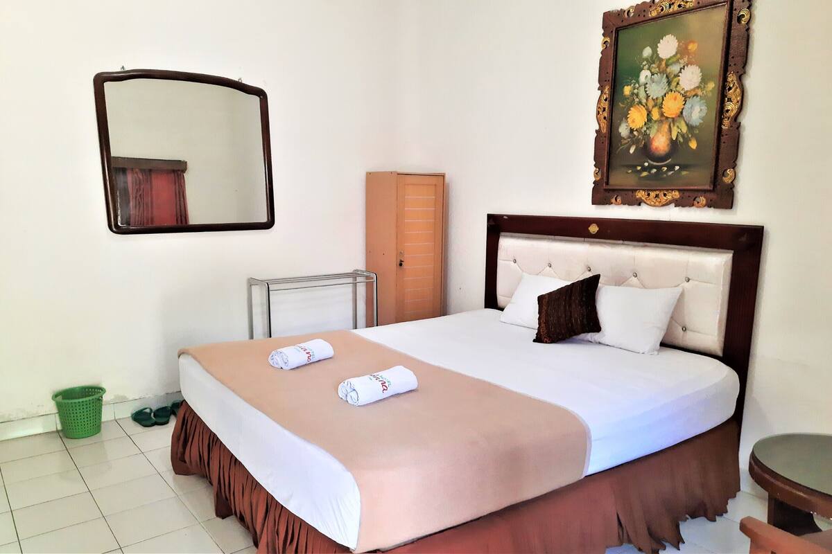 A hotel room with a bed, a mirror, a framed painting, a radiator, a green basket, and a door.