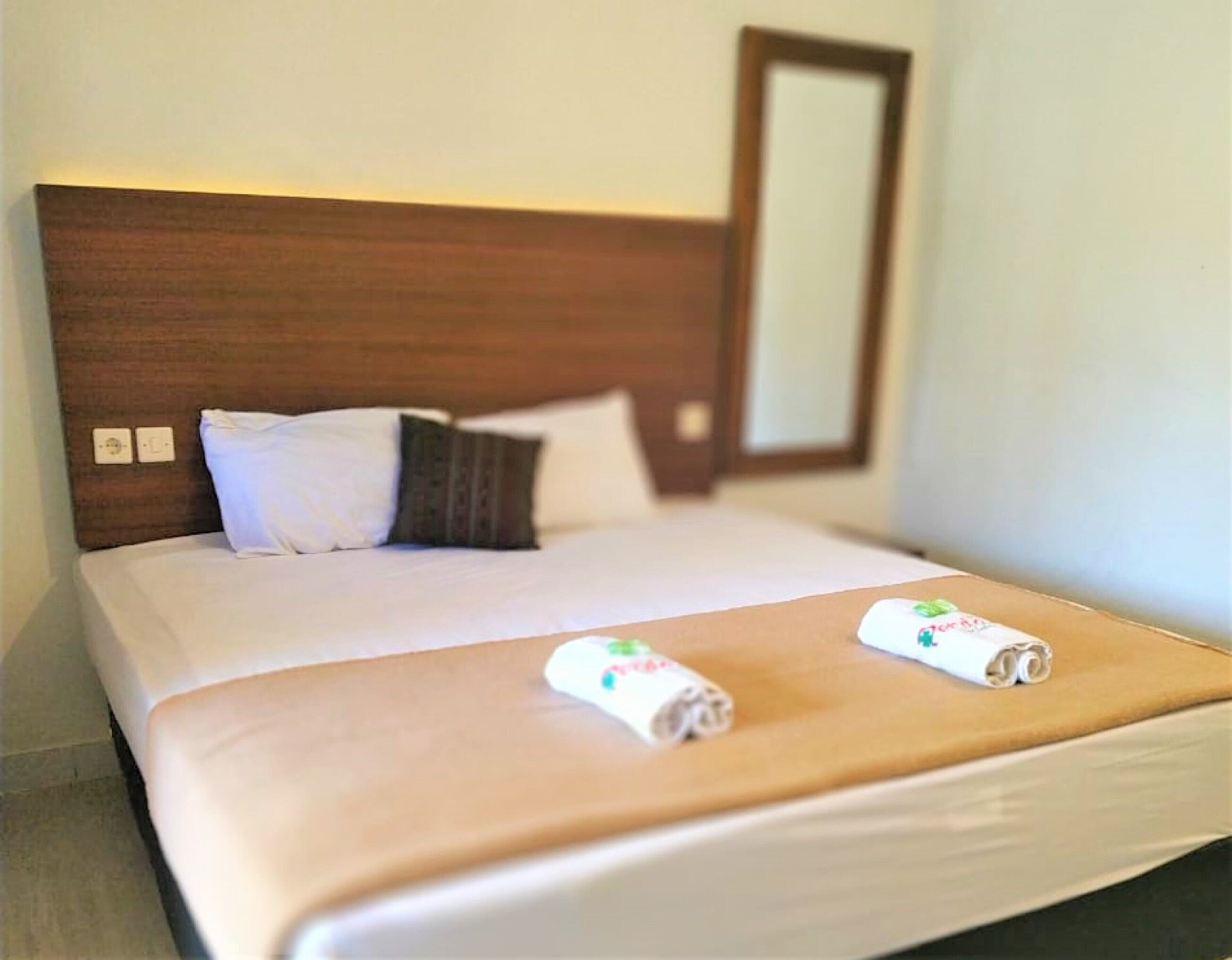 A neatly made bed with a brown headboard, white bedding, and two rolled towels with a green leaf design.