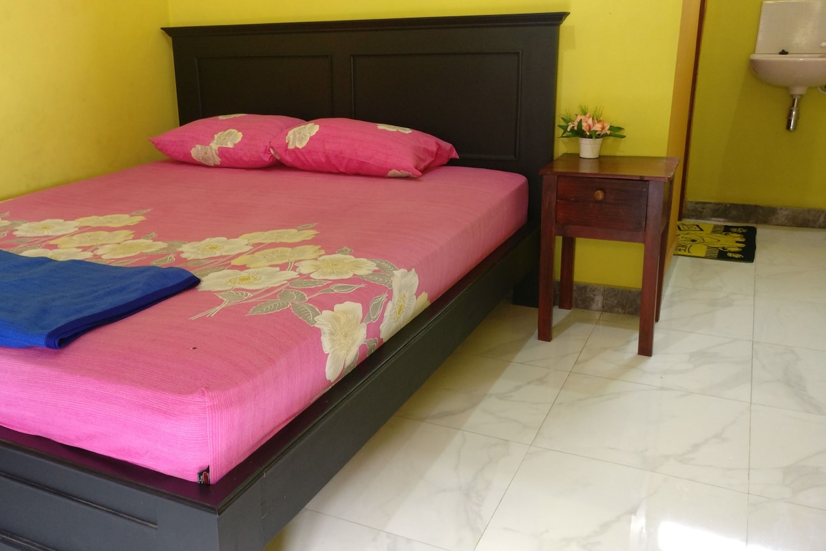 A bed with a floral bedspread and two pillows, a small wooden nightstand, a white sink, and a tiled floor.