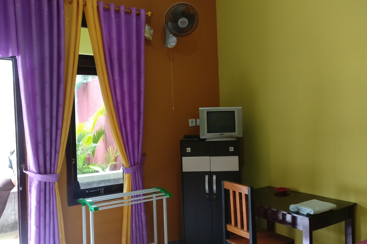 A room with a window covered by purple and yellow curtains, a television on a cabinet, a chair, and a table with a towel on it.