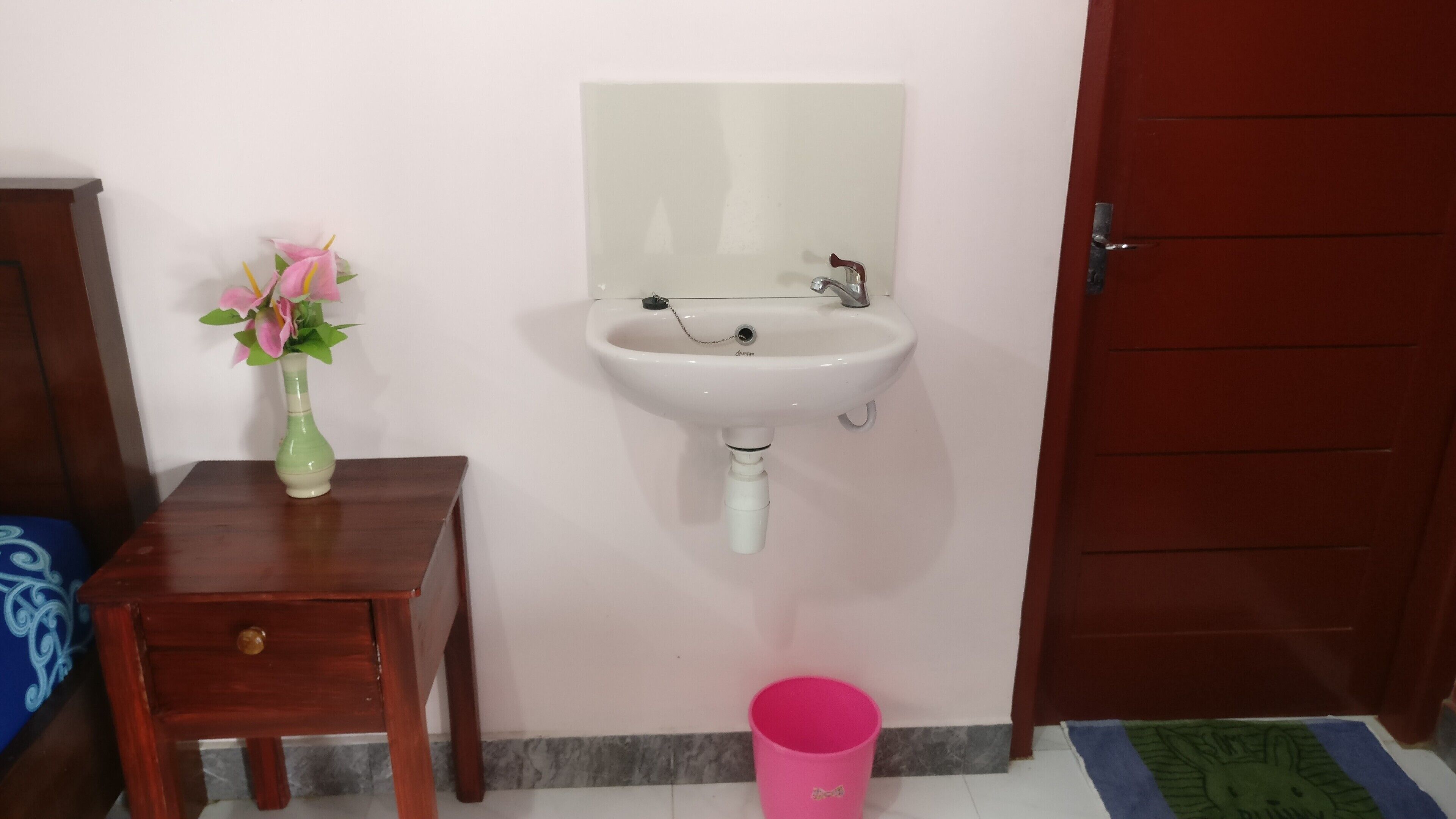 Une salle de bain avec un lavabo blanc, une petite table en bois sur laquelle se trouve un vase, et un seau rose posé sur le sol.