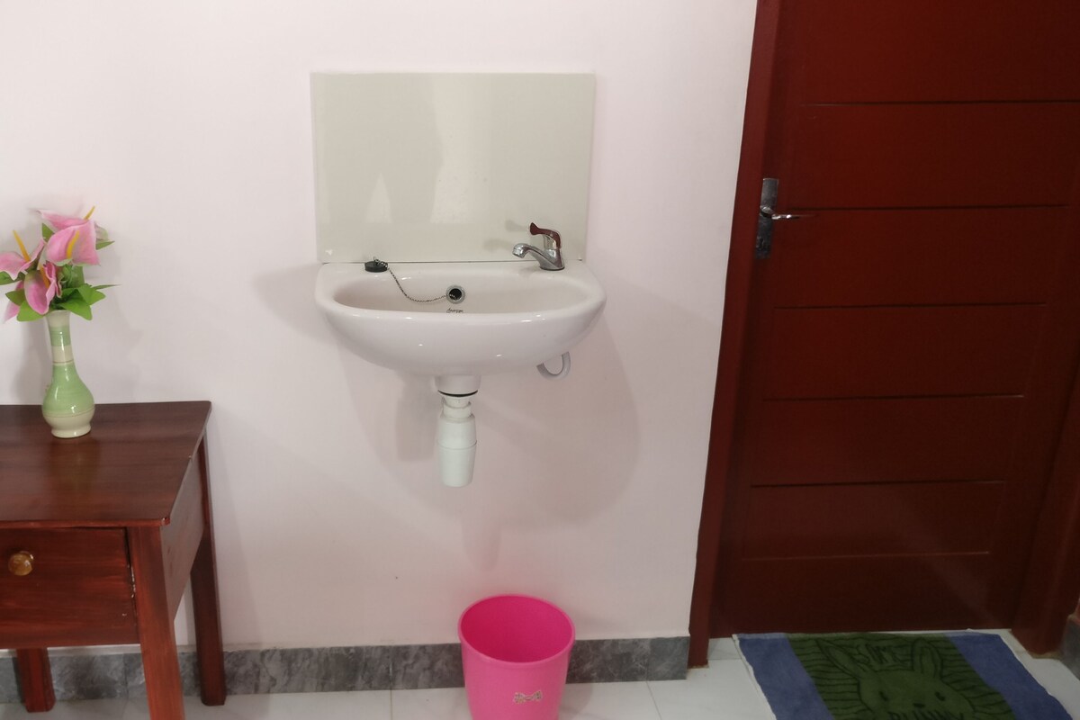 A bathroom with a white sink, a wooden side table with a vase of flowers, a pink trash bin, and a red door.