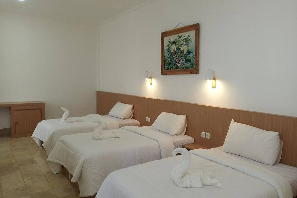 A hotel room with three beds, each with a white bedspread and matching pillows. There is a wooden headboard, a small bedside table, and a wall-mounted light fixture.