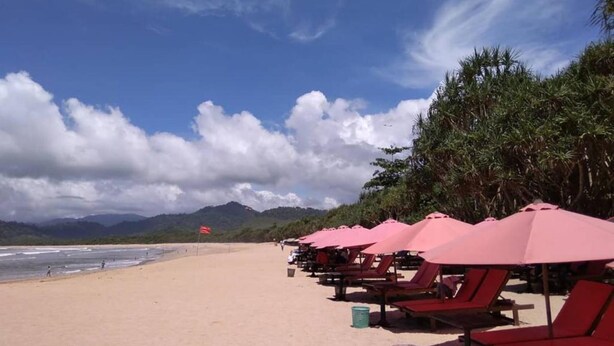 Ein Strand mit Reihen roter Liegen und Sonnenschirme, ein sandiger Uferbereich und ein Gebirge im Hintergrund.