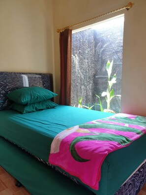 A bed with a teal bedspread and a colorful blanket, a green pillow, and a window with a view of a plant and a textured wall. - Nita's Homestay Banyuwangi (Banyuwangi)