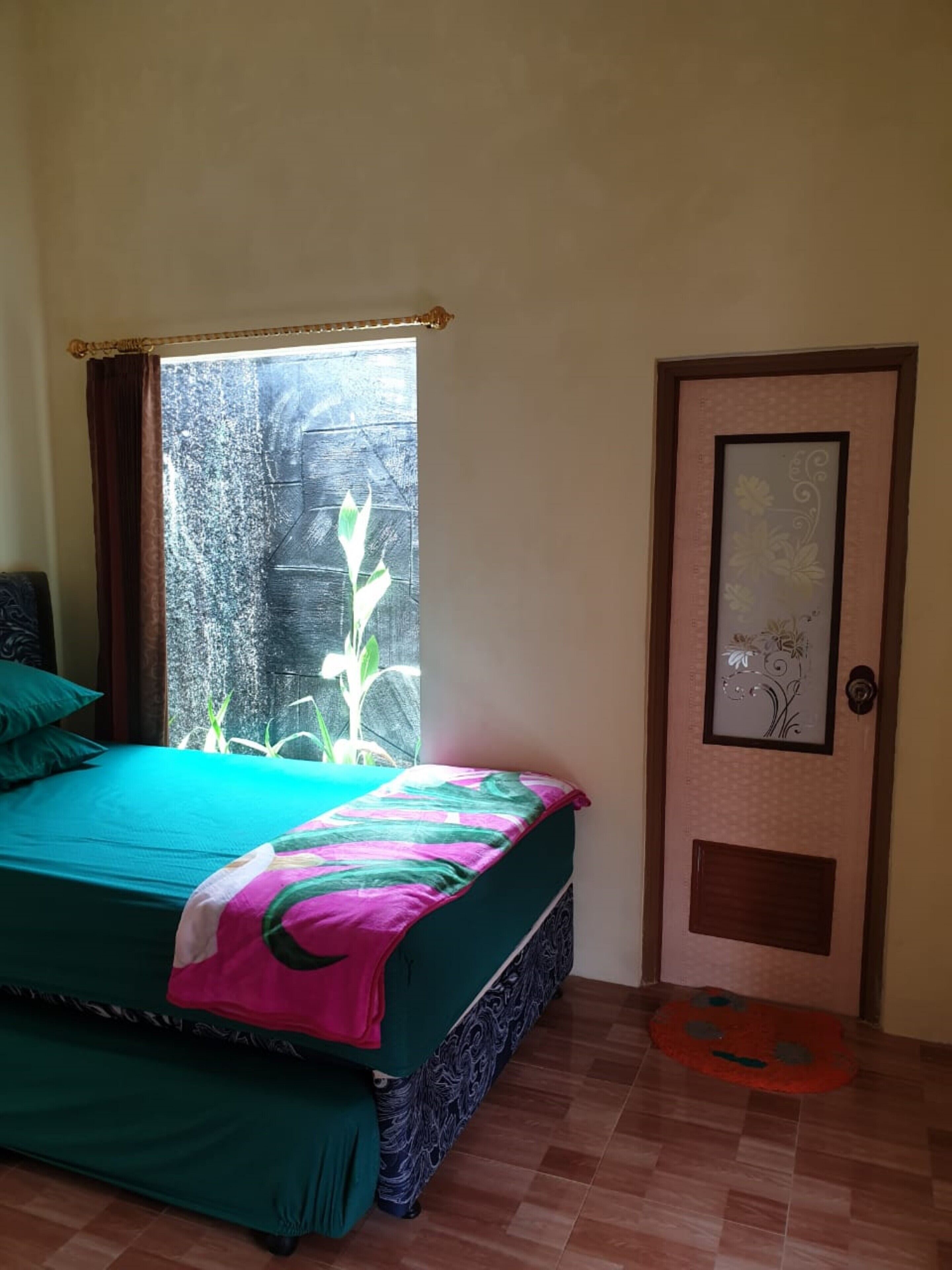 A bedroom with a bed, a window with a view of a plant, and a door with a decorative panel.