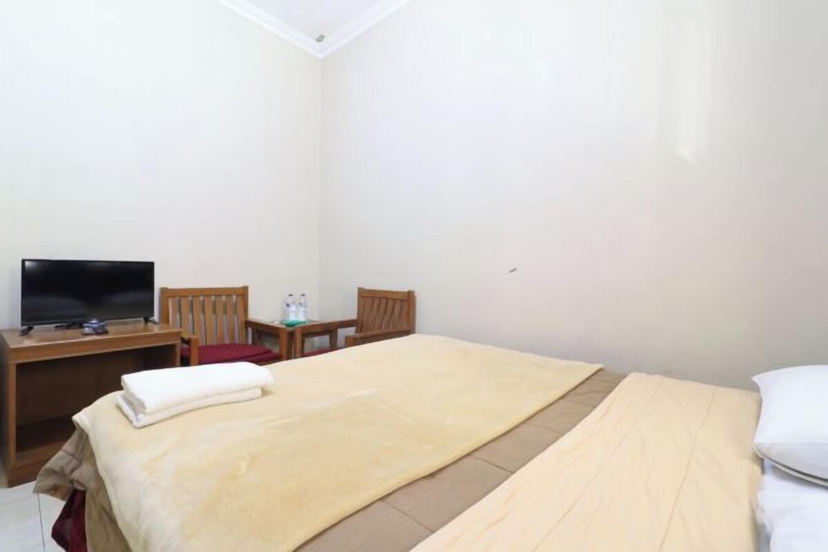 A hotel room with a bed, a desk with a television, two chairs, and a small table with a lamp.