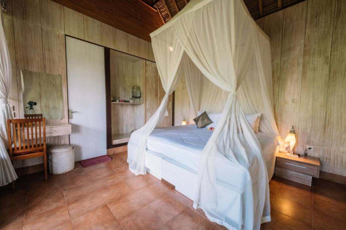 Een slaapkamer met een hemelbed, een houten stoel, een nachtkastje en een spiegel.