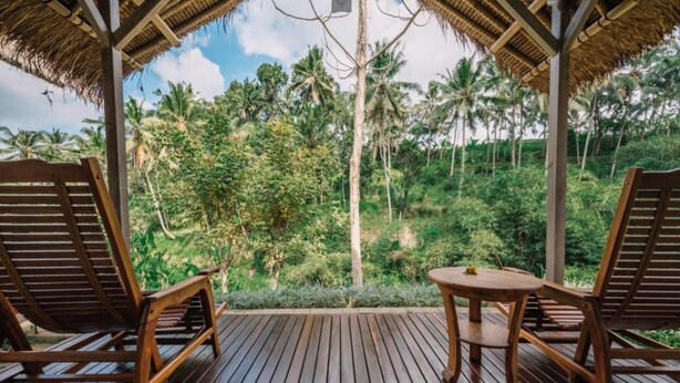 Een houten terras met twee loungesteunen en een bijzettafeltje, met uitzicht op een weelderig tropisch landschap met palmbomen.