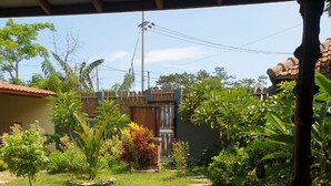 Um jardim com um caminho de pedra que leva a um portão de madeira, rodeado por diversas plantas e um espaço de pátio com azulejos.
