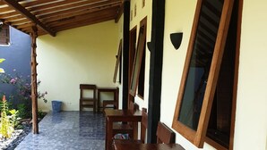 Um alpendre coberto com mesas e cadeiras de madeira, chão de azulejos e uma parede amarela com janelas.