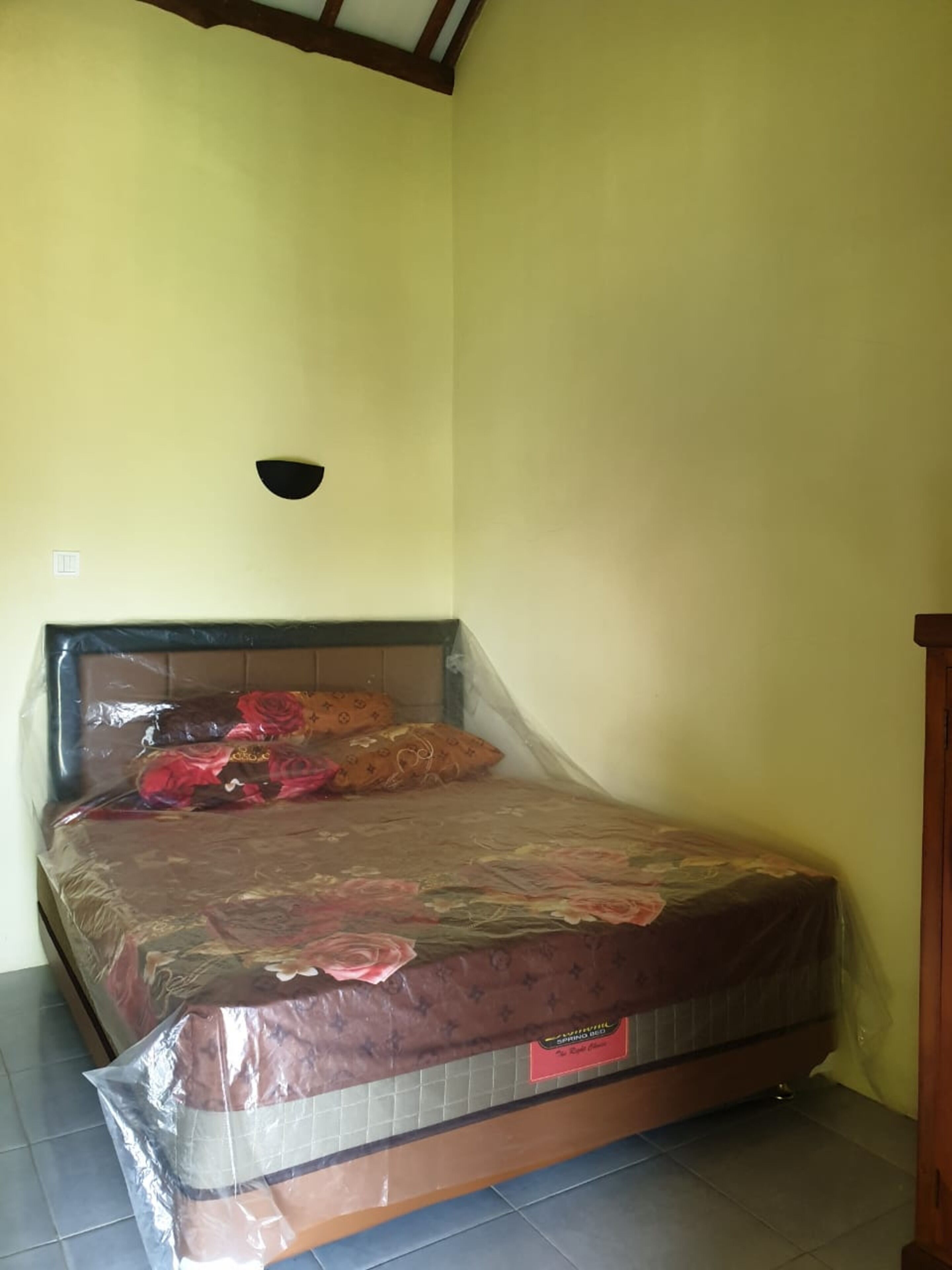 A room with a bed covered in a floral-patterned mattress protector, a dark headboard, and a wall-mounted light fixture.