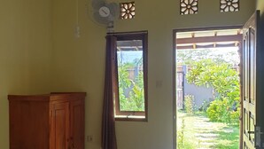 Quarto com cama, guarda-roupa de madeira, janela com cortinas, ventoinha e vista para um jardim através de uma porta aberta.