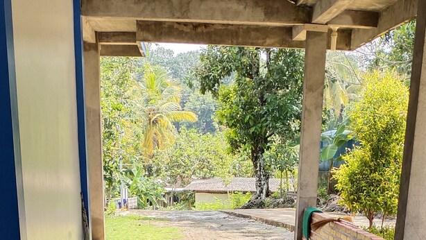 A covered outdoor area with a tiled floor, a brick wall, and a view of a garden with trees and a house in the background. - Homestay Sudiono (Mangunan)