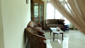 A room with a wooden cabinet, two upholstered chairs, a small table with a jar, and a window with sheer curtains. - Homestay Sudiono (Mangunan)