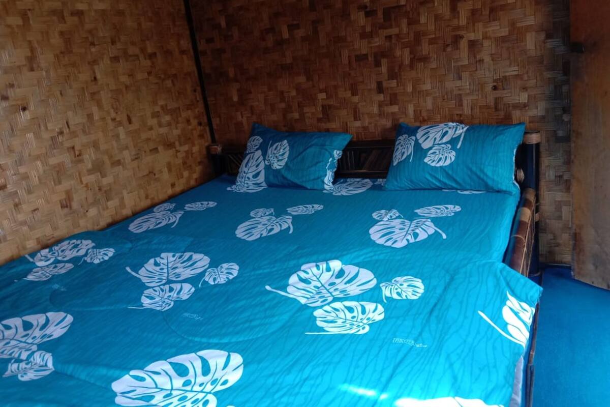 Una stanza con un letto ricoperto da biancheria azzurra con motivi a foglia bianca, una testiera di legno e una parete intrecciata.