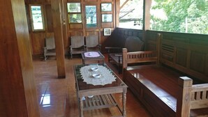 A wooden interior with a dining area featuring a table with a lace tablecloth and two metal plates, surrounded by wooden chairs and benches. - Homestay Arya Mangunan (Mangunan)
