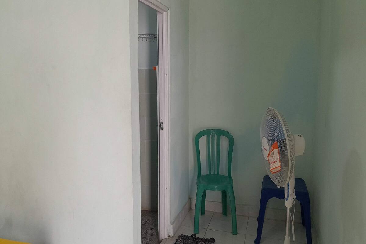 Ein Zimmer mit einer Tür zu einem Badezimmer, einem grünen Plastikstuhl und einem Standventilator auf einem blauen Hocker.