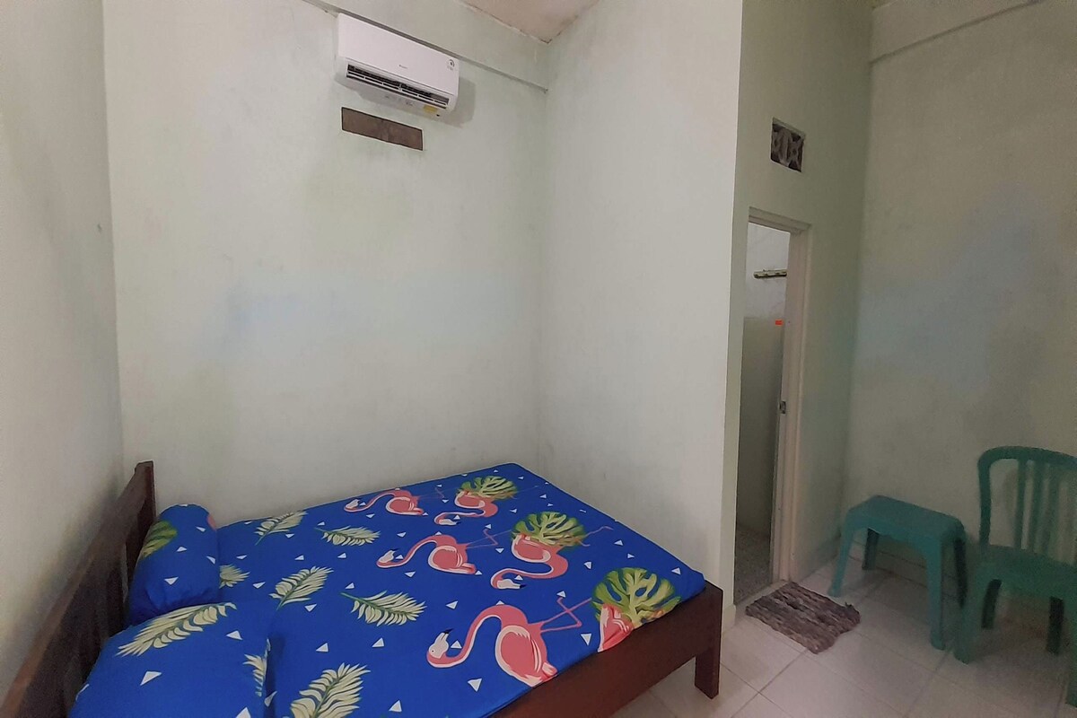 Ein kleines Einzelzimmer mit einem Bett, dessen blaue Bettdecke ein tropisches Blattmuster zeigt. Ein Holz-Kopfteil, ein weißes an der Wand montiertes Klimagerät und ein kleiner grüner Hocker in der Nähe der Tür sind ebenfalls vorhanden.
