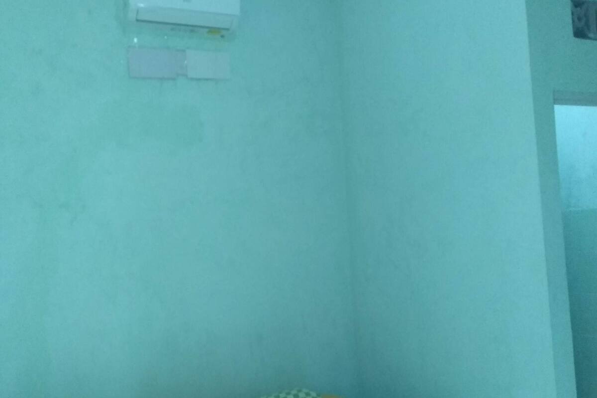 Ein Zimmer mit einer hellblauen Wand, einem kleinen Bett mit einem gemusterten Überwurf und einer weißen Wandklimaanlage.