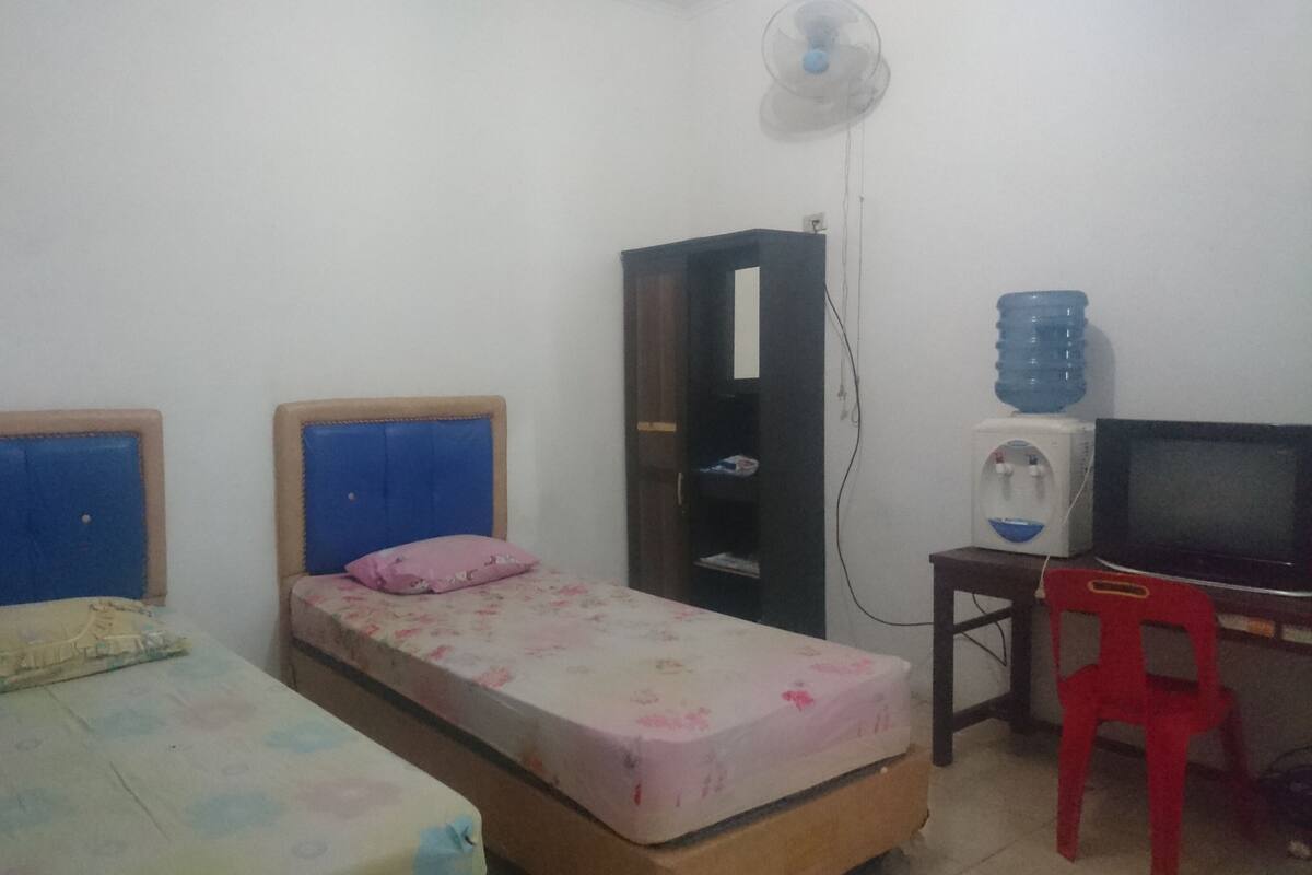 Una habitación pequeña con dos camas individuales, un escritorio con ordenador, dispensador de agua y una silla roja.