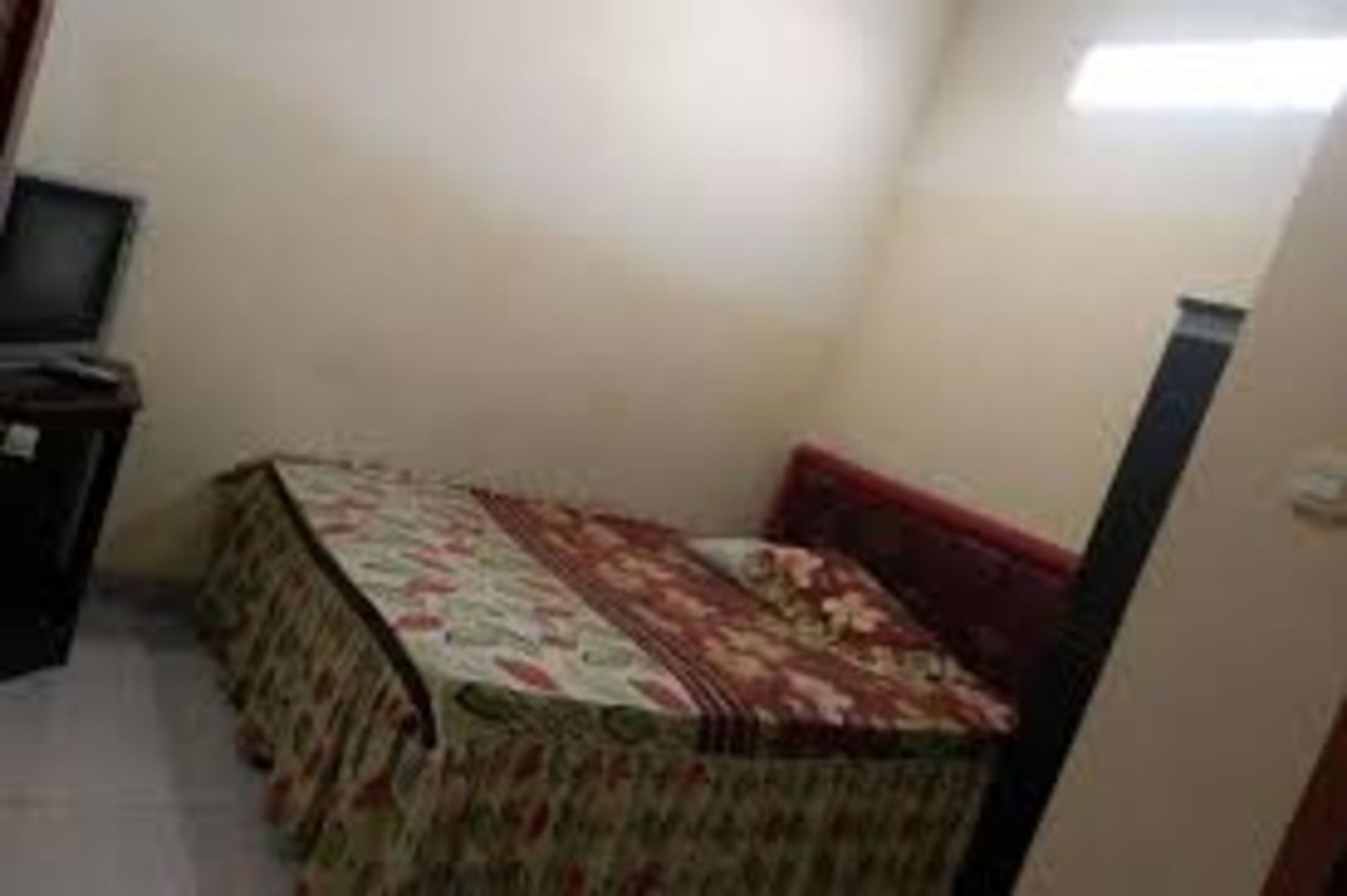 Een kleine, smalle kamer met een bed, overdekt met een bloemenprintdeedbed, een klein tafeltje met een lamp en een televisie op een standaard.