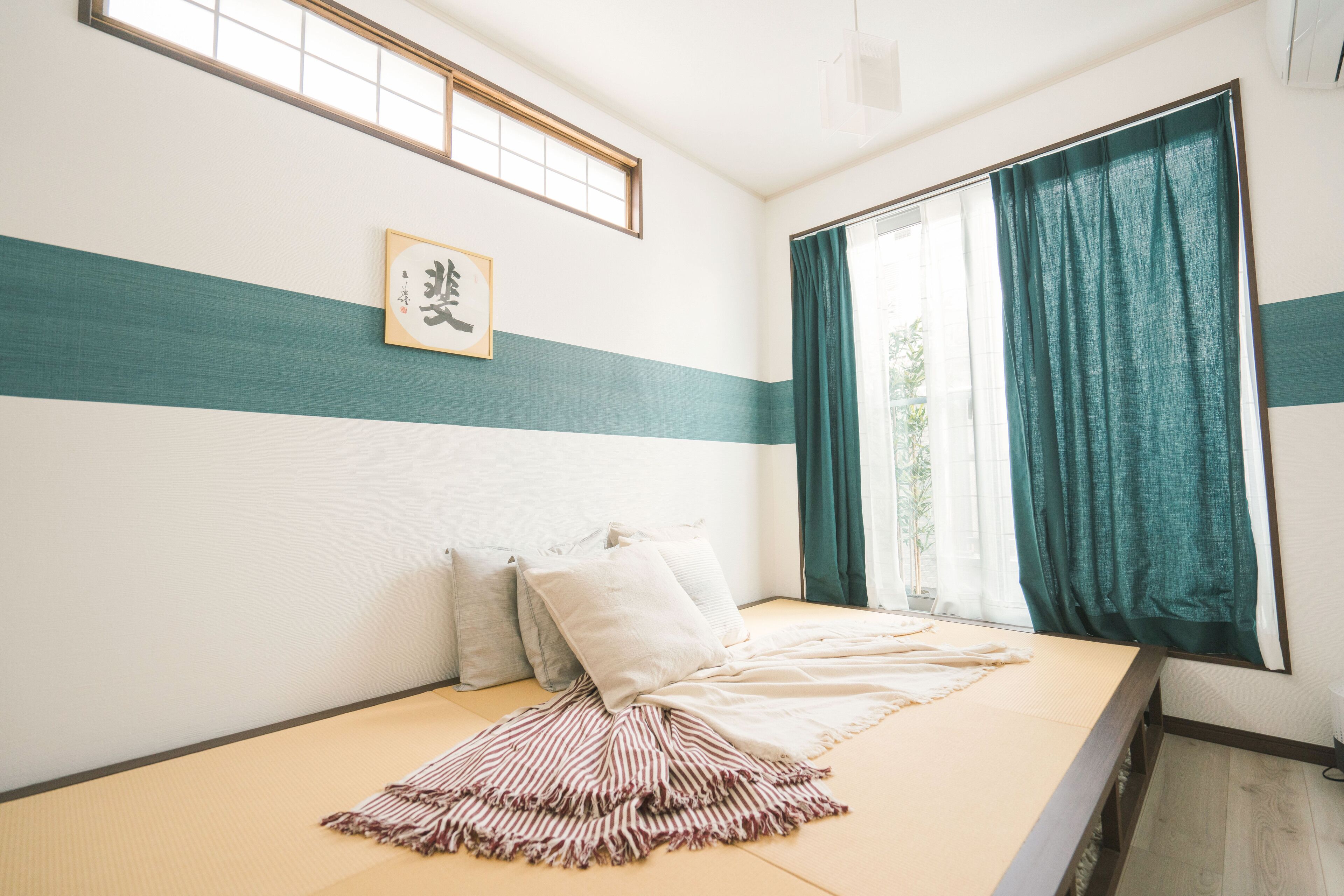 A room with tatami flooring, a bed with pillows, a framed picture on the wall, and a window with curtains.