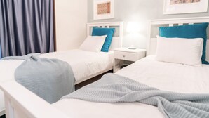 A hotel room with two beds, each with a white and blue pillow, a nightstand with a lamp, and a framed picture on the wall. - Back to 90s American Style House (Tokyo)