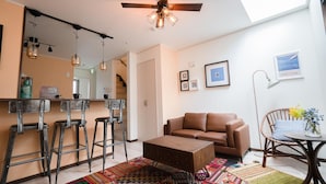 A modern living room with a brown leather sofa, a wooden coffee table, and a colorful rug. There are two bar stools, a dining table with chairs, and a ceiling fan. - Back to 90s American Style House (Tokyo)