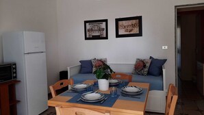 Ein kompakter Wohnbereich mit Esstisch für zwei Personen, einer Couch mit Kissen, einem weißen Kühlschrank und zwei gerahmten Bildern an der Wand.