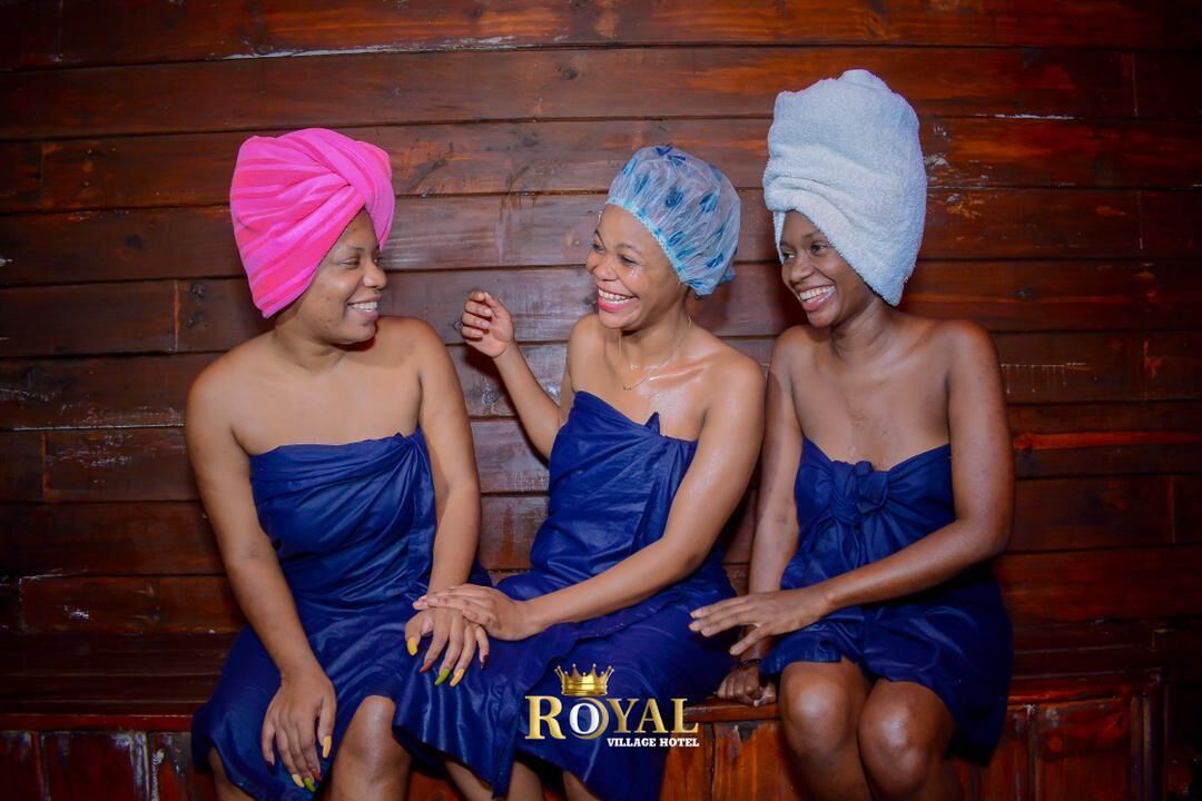 Three individuals seated on a bench in a wooden cabin setting, wearing blue garments and hair wraps.