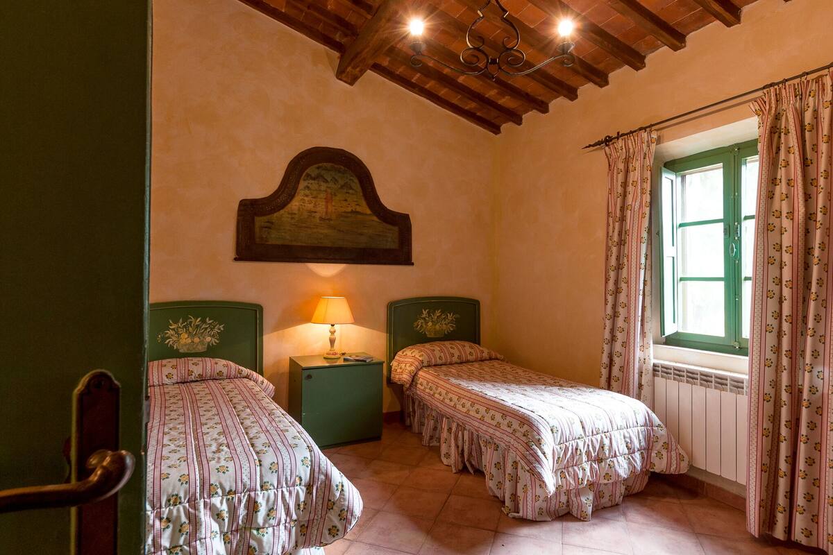 Ein Zimmer mit zwei Einzelbetten, einem Nachttisch mit Lampe, einer Holzbalkendecke und einem Fenster mit Vorhängen.