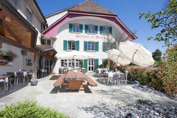 A traditional building with a sign reading "Gaißhof zum Bären". It features outdoor seating with tables and chairs, a stone-paved patio, and a large umbrella.