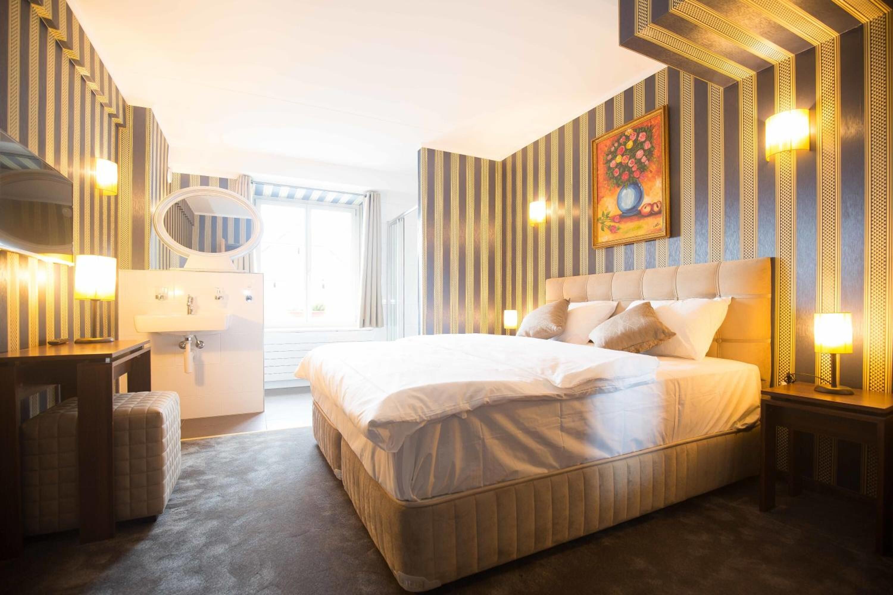 Ein Hotelzimmer mit einem großen Bett, einem Schreibtisch mit Stuhl, einem Schminktisch mit Spiegel und einem Bild an der Wand.
