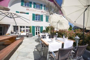 An outdoor dining area with tables and chairs, umbrellas, and a building with green shutters and a sign.