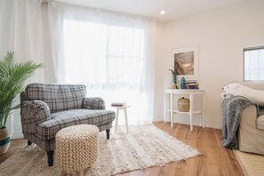 A cozy living room with a plaid sofa, a small ottoman, a white rug, a side table with books, and a potted plant.